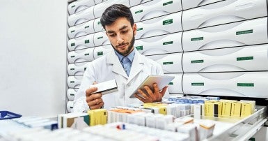 Confident male pharmacist reading label on merchandise in storage room. Young chemist is working in storage room. He is wearing lab coat at drugstore.